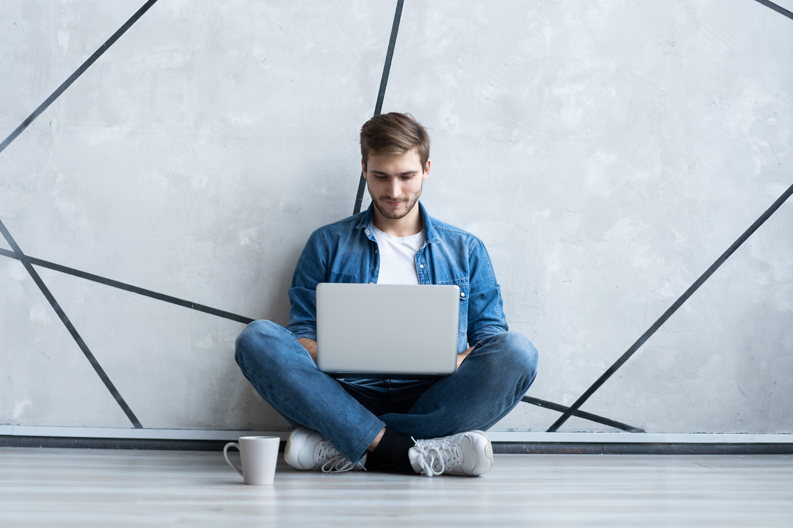 Young man working on laptop sitting on floor. Guy with computer reading news, surfing net or freelancing.